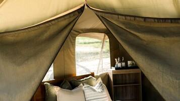 A tented accommodation with a bed, pillows, a small wooden shelf, and a window.