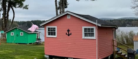 A small pink cabin with an anchor symbol, a green shed, and a lake in the background.