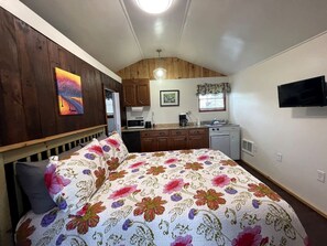 Quarto com cama, escrivaninha, cadeira, janela e um espaço de cozinha.