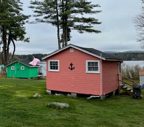 A small pink cabin with an anchor symbol, a green shed, and a lake in the background. - Lakefront Secluded Cottage in Northwood, New Hampshire (Northwood)