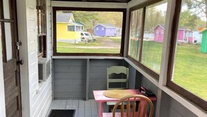 Varanda coberta com mesa e cadeira, janela com vista para casas e uma porta.