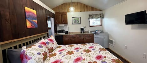 A bedroom with a bed, a desk, a chair, a window, and a kitchen area.