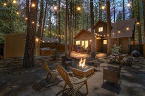 Eine Holzhütte mit Feuerstelle, Sitzgelegenheit im Freien und Lichterketten in einer Waldlandschaft.