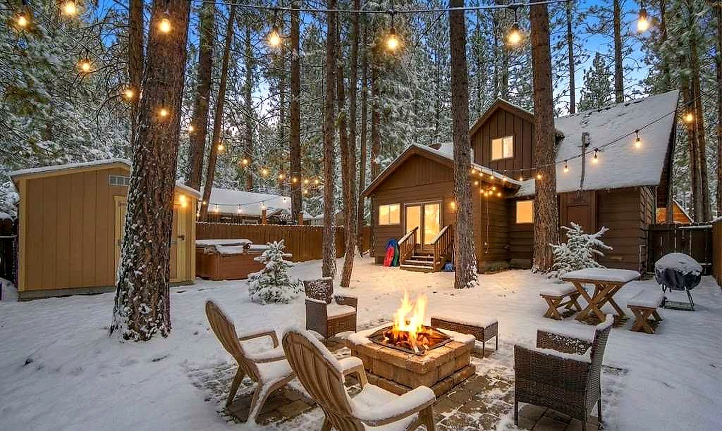 Een chalet met een vuurkuil, buitenmeubels en lichtslingers, gelegen in een besneeuwd bos.