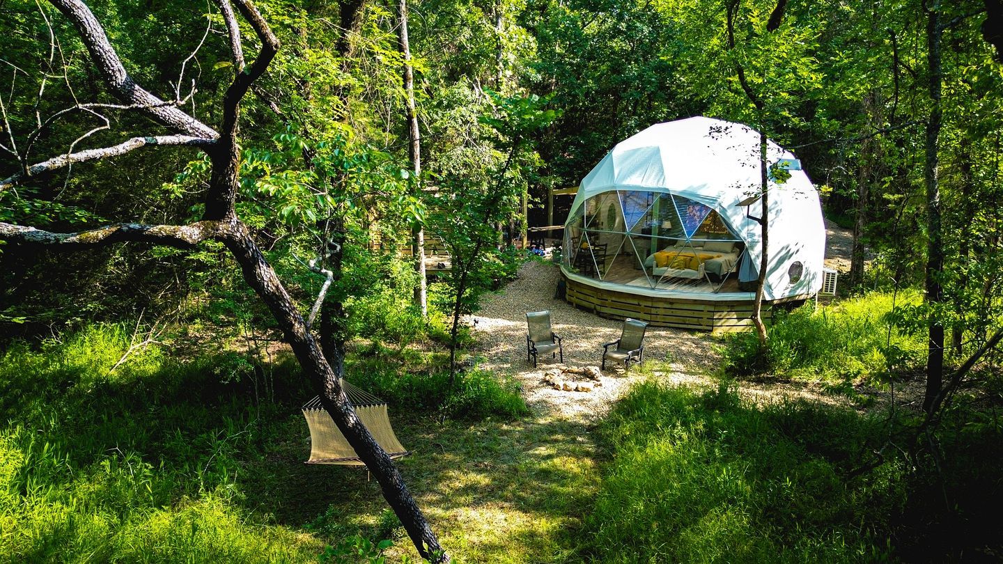 A dome-shaped tent with a bed and seating area, surrounded by trees and a gravel path.