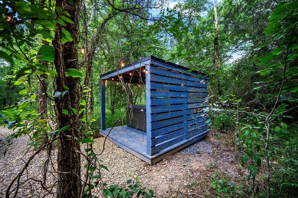 A small, blue wooden structure with a slatted roof, surrounded by dense forest.