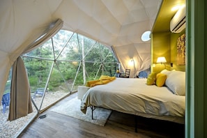 A tented room with a bed, a chair, a lamp, and a view of the forest.
