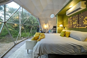A modern tent-style room with a large bed, a seating area, and a view of the forest.