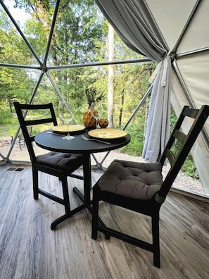 Un coin repas avec une table et des chaises, un vase et une bouteille, le tout mis en valeur par un décor d’arbres en arrière-plan.