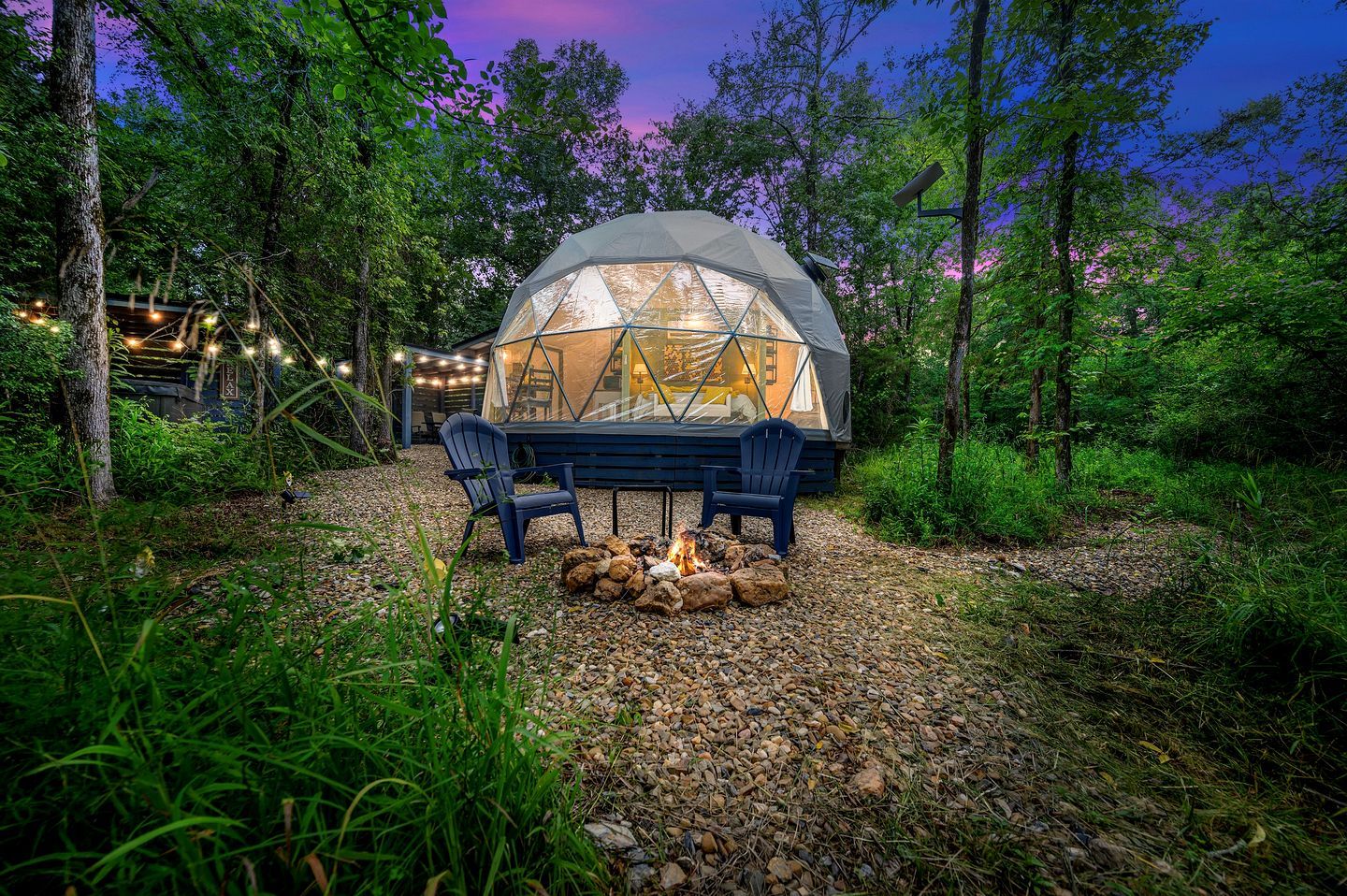 A geodesic dome structure with a fire pit and two chairs in a forested area.
