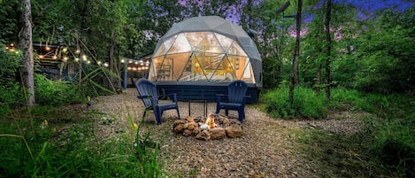 A geodesic dome structure with a fire pit and two chairs in a forested area.