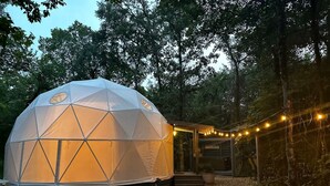 Una estructura de cúpula geodèsica amb un camí de grava, envoltada d'arbres i un edifici de fusta amb un rètol.