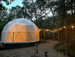 A geodesic dome structure with a gravel path, surrounded by trees and a wooden building with a sign.