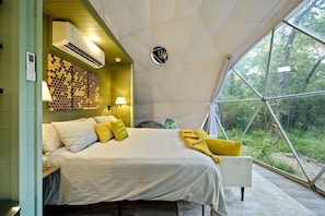 A bedroom with a bed, pillows, a wall-mounted air conditioner, a lamp, and a view of the outdoors through a large window.
