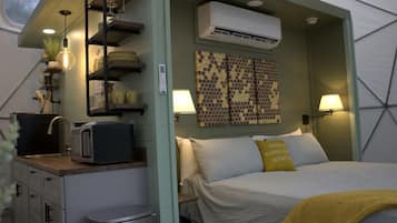 A compact room with a bed, bedside lamps, a small table with a microwave, a trash bin, and a wall-mounted air conditioner.