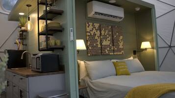 Habitación compacta con cama, lámparas de noche, una pequeña mesa con microondas, un cubo de basura y aire acondicionado de pared.