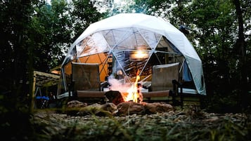A geodesic dome tent with a fire in front, surrounded by trees.