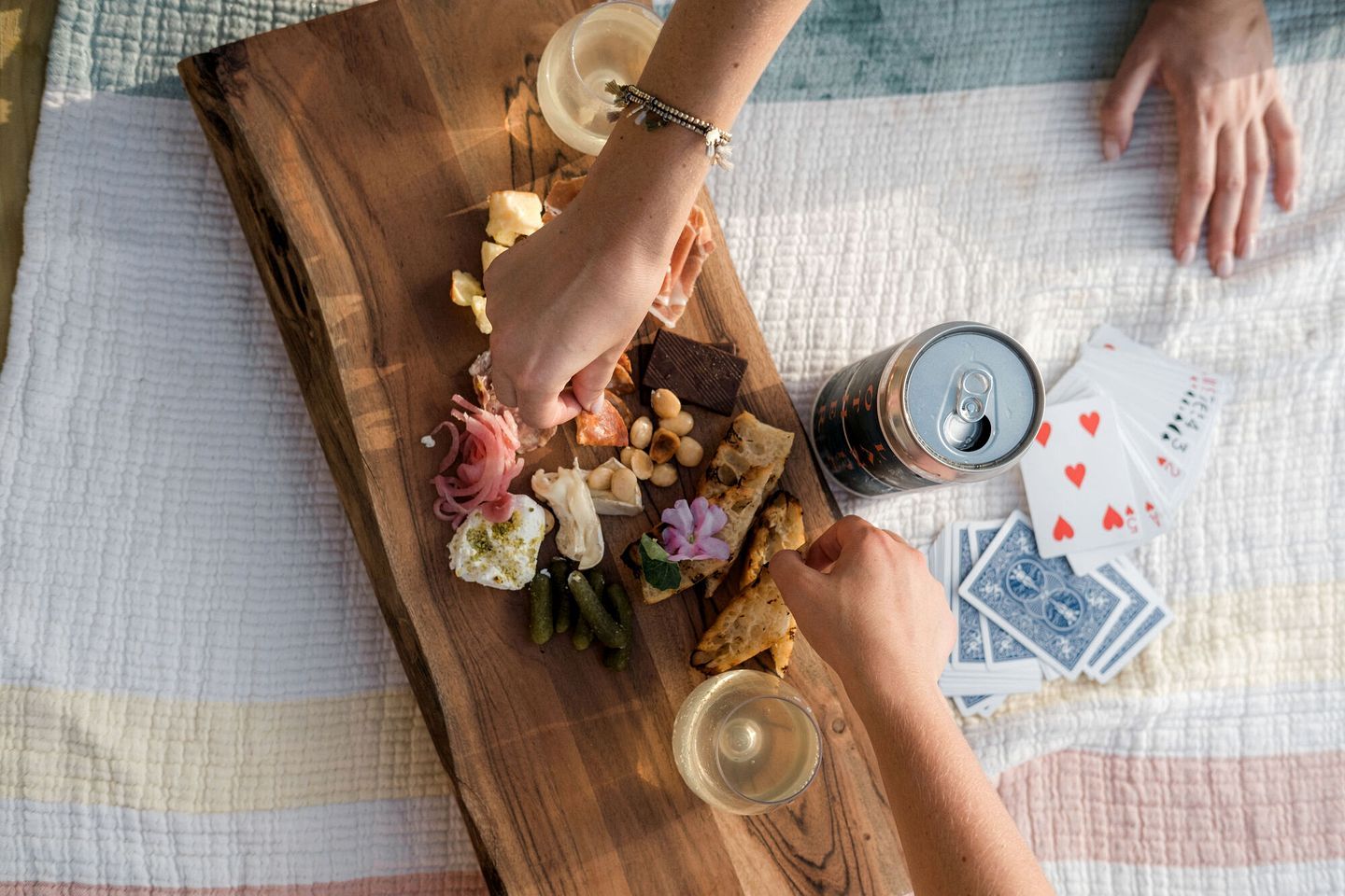 A wooden board with various food items and a can of beer, a glass of wine, and playing cards on a table.
