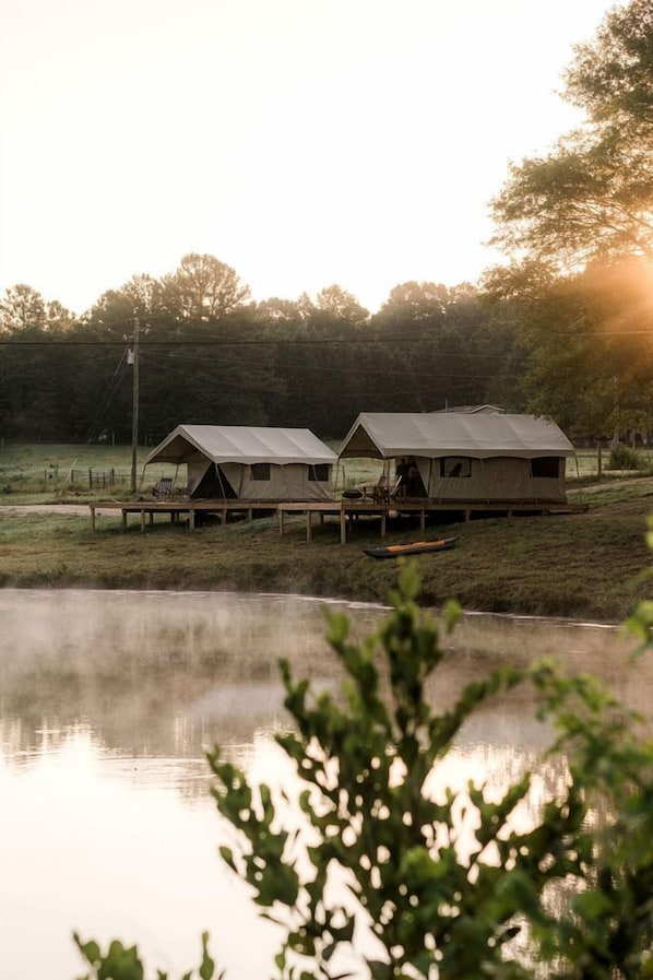 A row of tents with a wooden deck in front, a body of water in the foreground, and trees in the background.