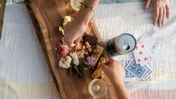 A wooden board with various food items and a can of beer, a glass of wine, and playing cards on a table.