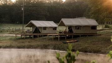 A row of tents with a wooden deck in front, a body of water in the foreground, and trees in the background.