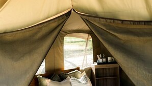 A tented accommodation with a bed, pillows, a small wooden shelf, and a window.