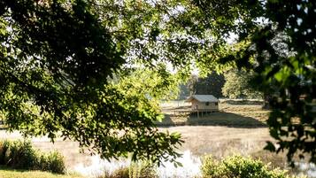 Une structure en forme de tente, située au bord d’un plan d’eau, entourée d’arbres et d’herbe.