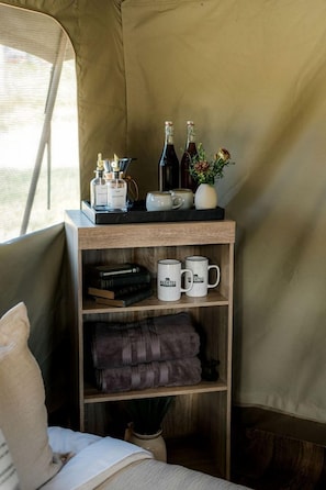 A tent interior with a shelf holding books, mugs, and bottles; a bed with a pillow; and a window with a view.