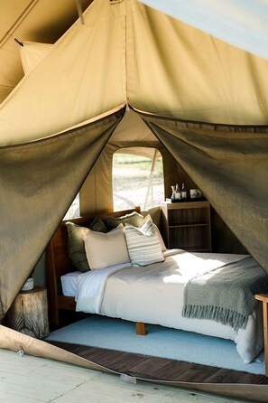 A tented accommodation with a bed, pillows, a small wooden shelf, and a window.