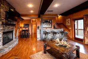A spacious living room with a stone fireplace, wooden flooring, a dining area with bar stools, a staircase, and a large window with curtains. - Lakeview Cabin with Large Private Deck in Topton, North Carolina (Topton)