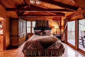 A wooden cabin bedroom with a bed, a nightstand, a dresser, and a window overlooking a deck.
