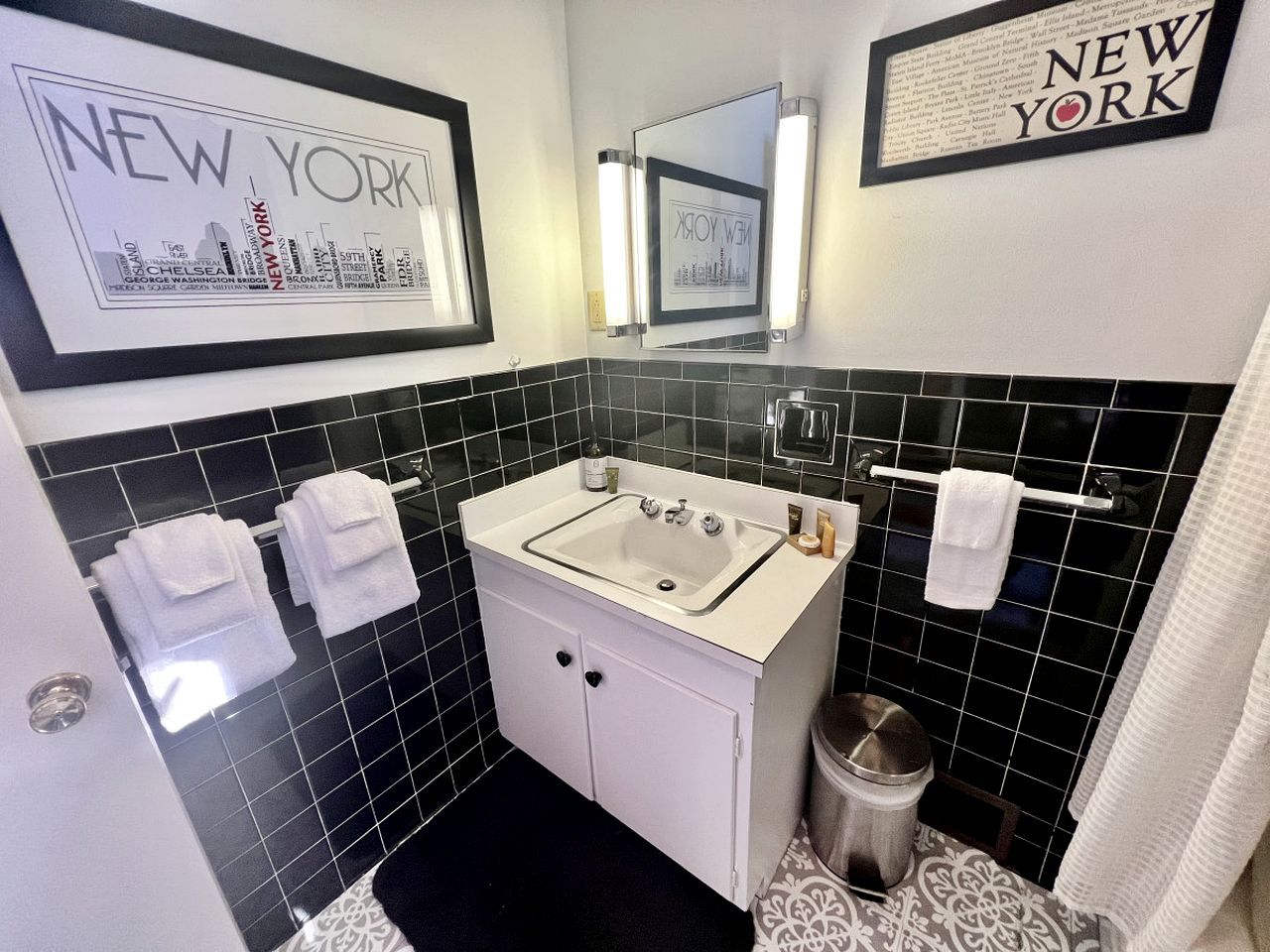 Ein Badezimmer mit schwarz-weißen Fliesen, einem Waschbecken, einem Spiegel und gerahmten Bildern von New York.
