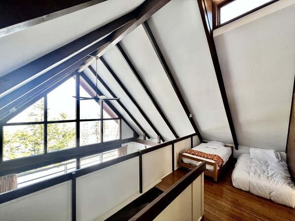 A room with a sloped ceiling, wooden flooring, a bed, a small table, and a window with a view of trees.