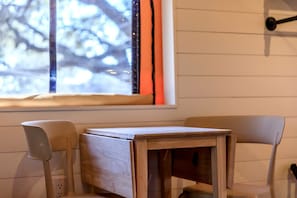 Une petite table en bois avec deux chaises, une fenêtre donnant sur des arbres et un luminaire fixé au mur.