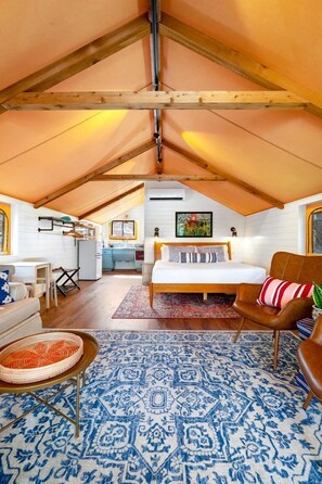 A cozy attic bedroom with a bed, a chair, a small table, and a kitchen area in the background.