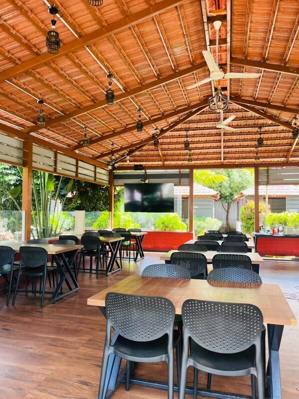 An outdoor dining area with wooden tables and chairs, a TV screen, and a thatched roof. - HOTEL KABAB JUNCTION (Bengaluru)