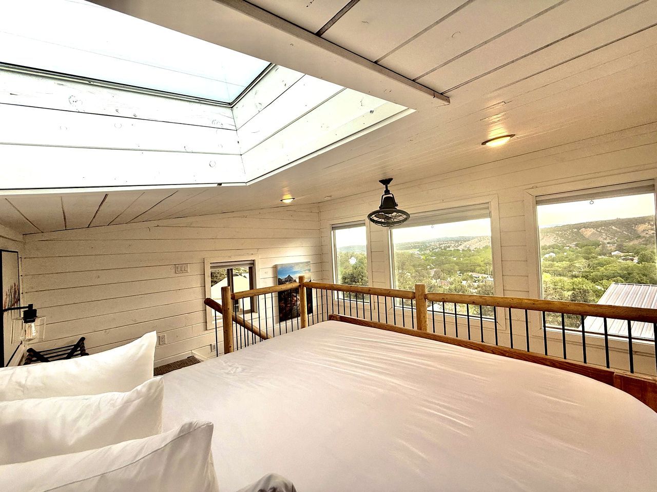 Une chambre avec une lucarne, un lit avec des oreillers, une balustrade en bois et une vue sur le paysage.