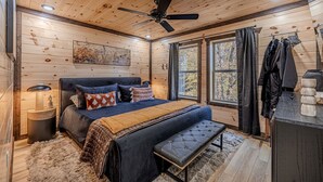 Un dormitorio con paneles de madera, una cama, un banco, un ventilador de techo y vistas a árboles a través de la ventana.