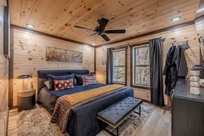 Ein Schlafzimmer mit Holzwandverkleidung, einem Bett, einer Bank, einem Deckenventilator und Blick auf Bäume durch das Fenster.