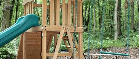 Une structure de jeux en bois, avec un toboggan et des balançoires, située dans un espace boisé.