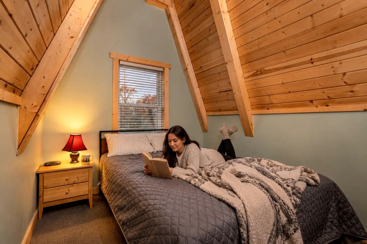 Un dormitorio con techo inclinado, una cama con colcha acolchada, una mesita de noche con lámpara y una ventana con persianas.