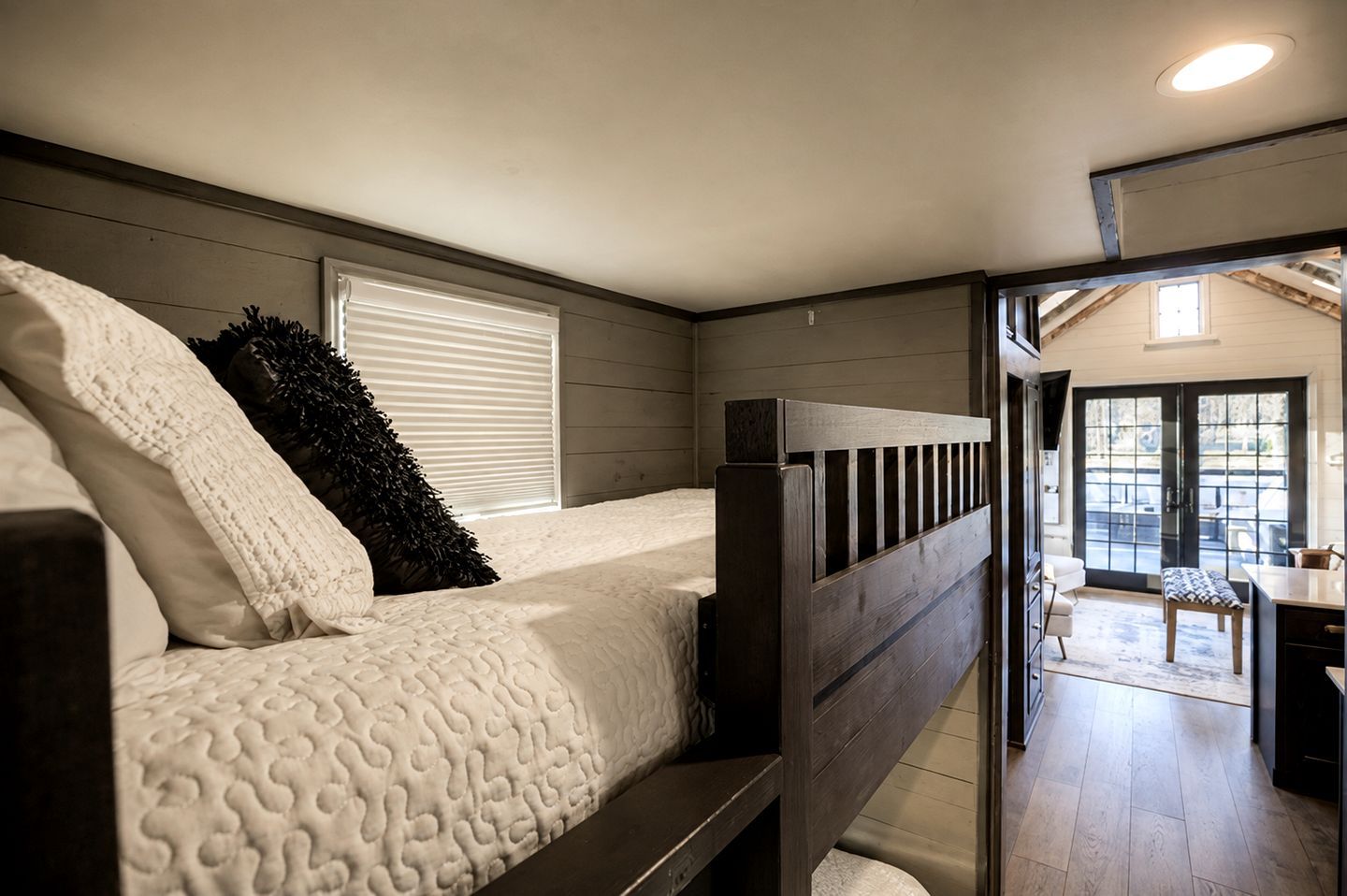 A bedroom with a bunk bed, a window with blinds, and a view of a dining area.