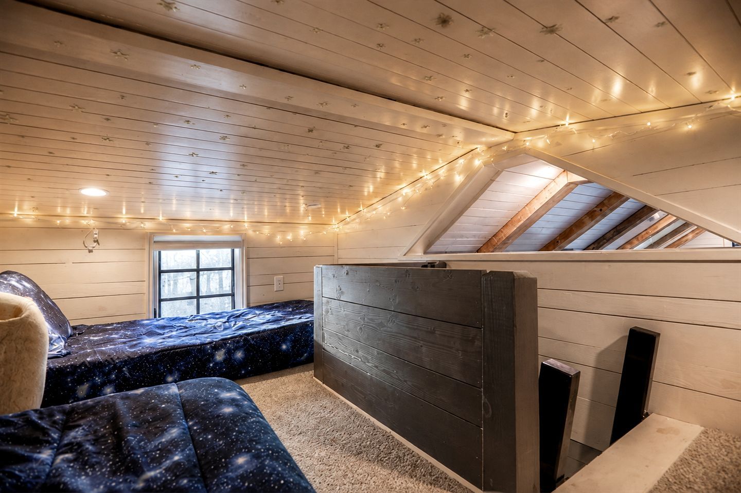 Een kamer met een schuine plafond, een bed met een sterrenhemel-dekbedovertrek, een houten hoofdbord en lichtslingers.