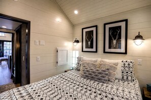 Un dormitorio con cama, dos cuadros enmarcados, una lámpara de pared y una puerta que conduce a otra habitación.