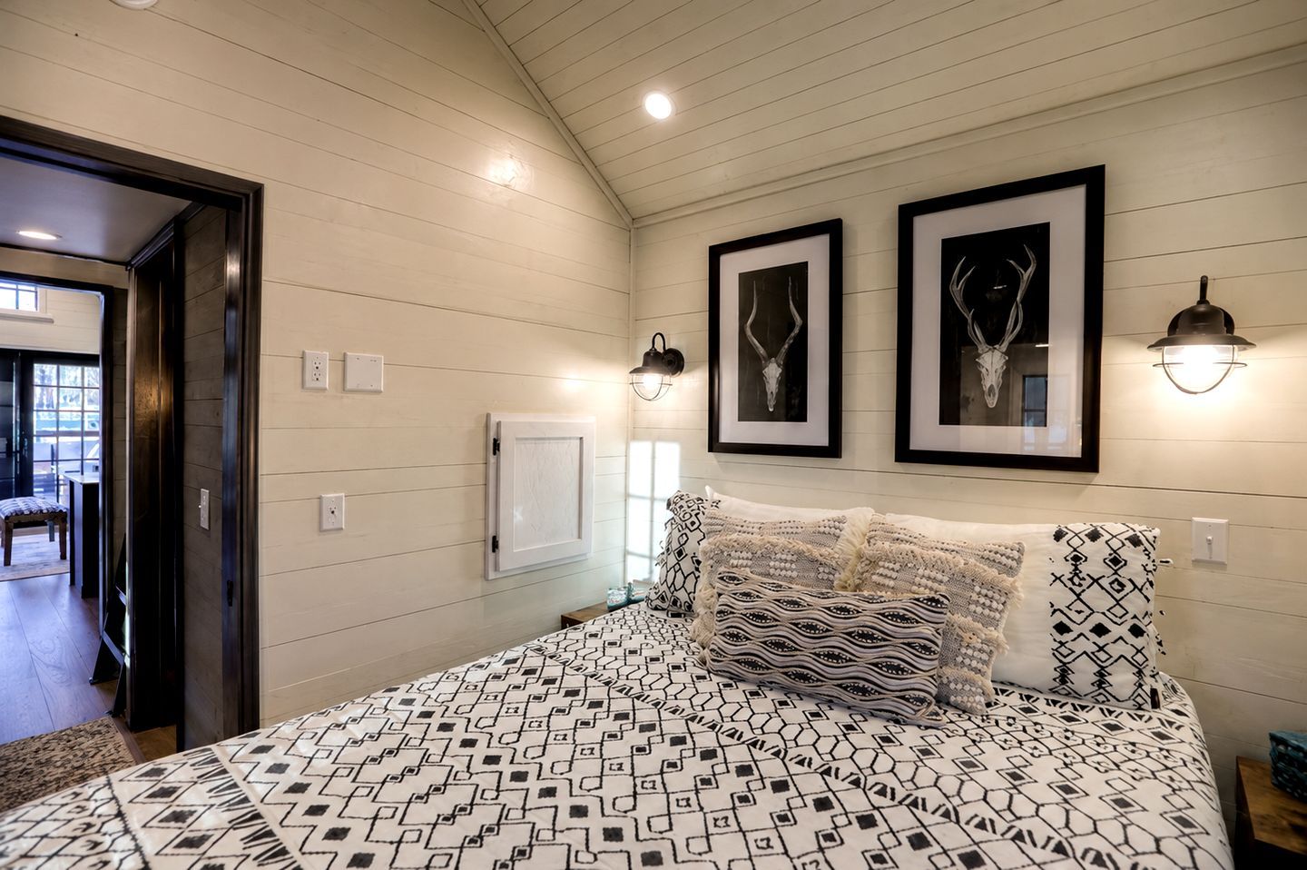 A bedroom with a bed, two framed pictures, a wall-mounted light, and a door leading to another room.