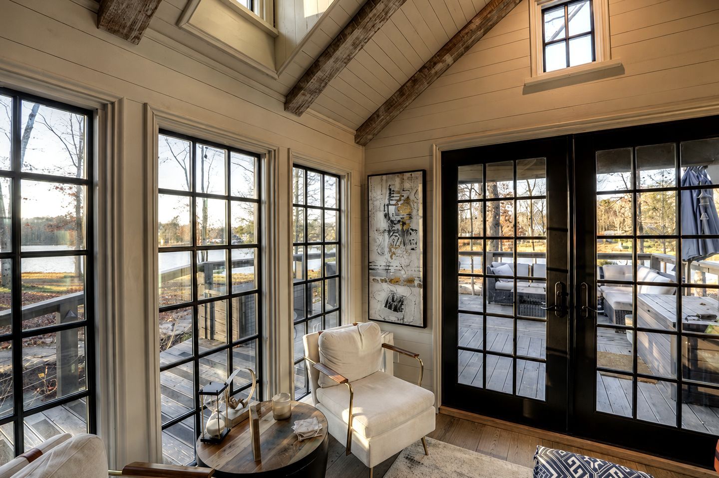 A room with large windows, a white armchair, a round table, and a view of a deck with outdoor furniture.