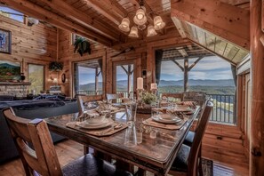 Une salle à manger en bois avec une grande table dressée pour quatre personnes, de grandes fenêtres offrant une vue sur les montagnes et un coin salon confortable avec une cheminée.