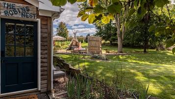 En rustik träbyggnad med en skylt som visar texten ”The Robin’s Nest”, en ytterdörr, en liten trädgård med en träbänk och en tipi-liknande konstruktion i bakgrunden.
