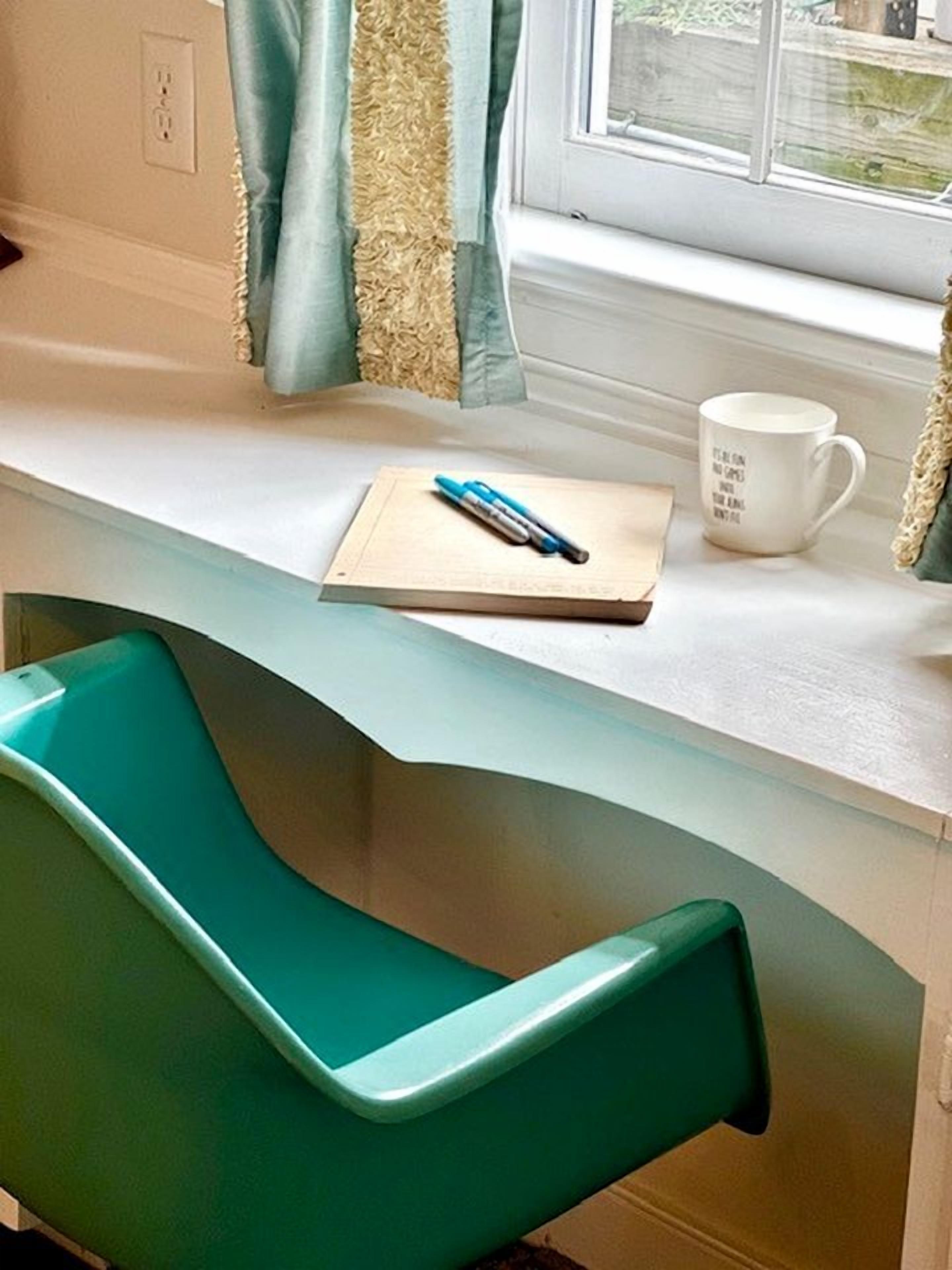 A room with a teal chair, a white desk, a notebook, a pen, and a mug.