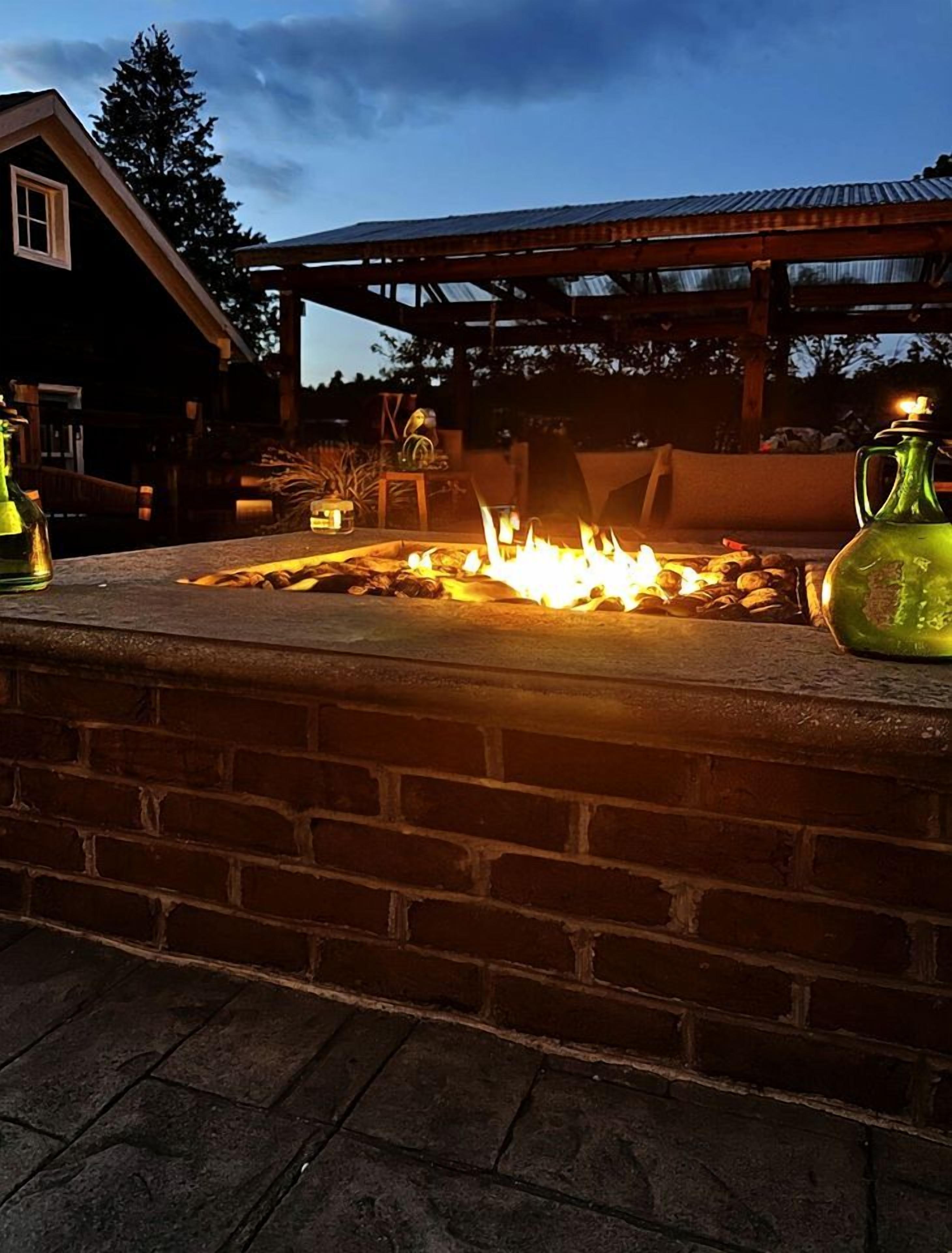 Bålsted med brænde, murstensvæg og en trækonstruktion i baggrunden.